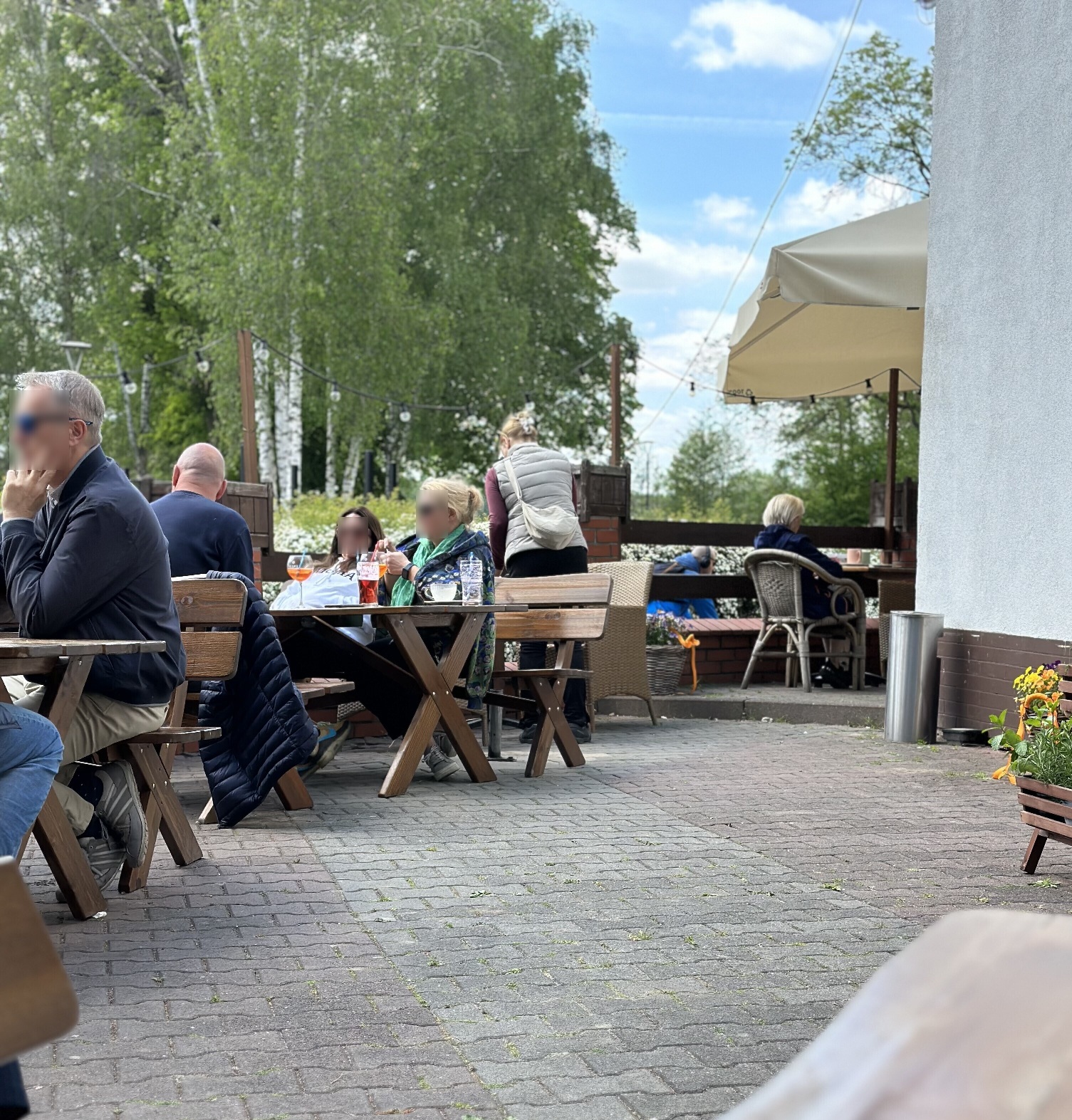 Zdjęcie z ogródka restauracji 'Przystań Smaków'. Drewniane stoły i ławy na kostce brukowej. W tle drzewa.