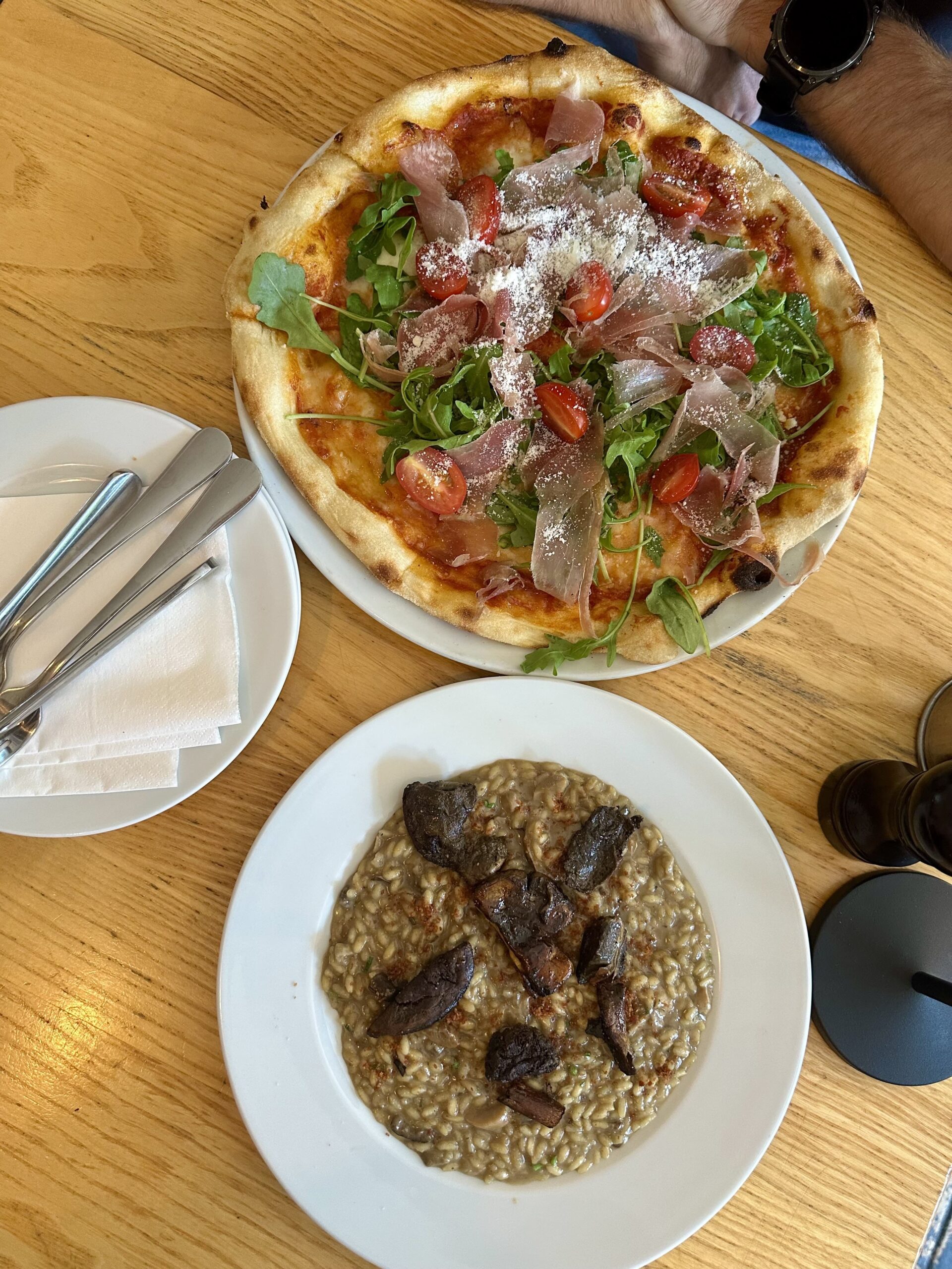 Risotto grzybowe ze smażonymi kawałkami grzybów podane na białym talerzu; w tle leży pizza.