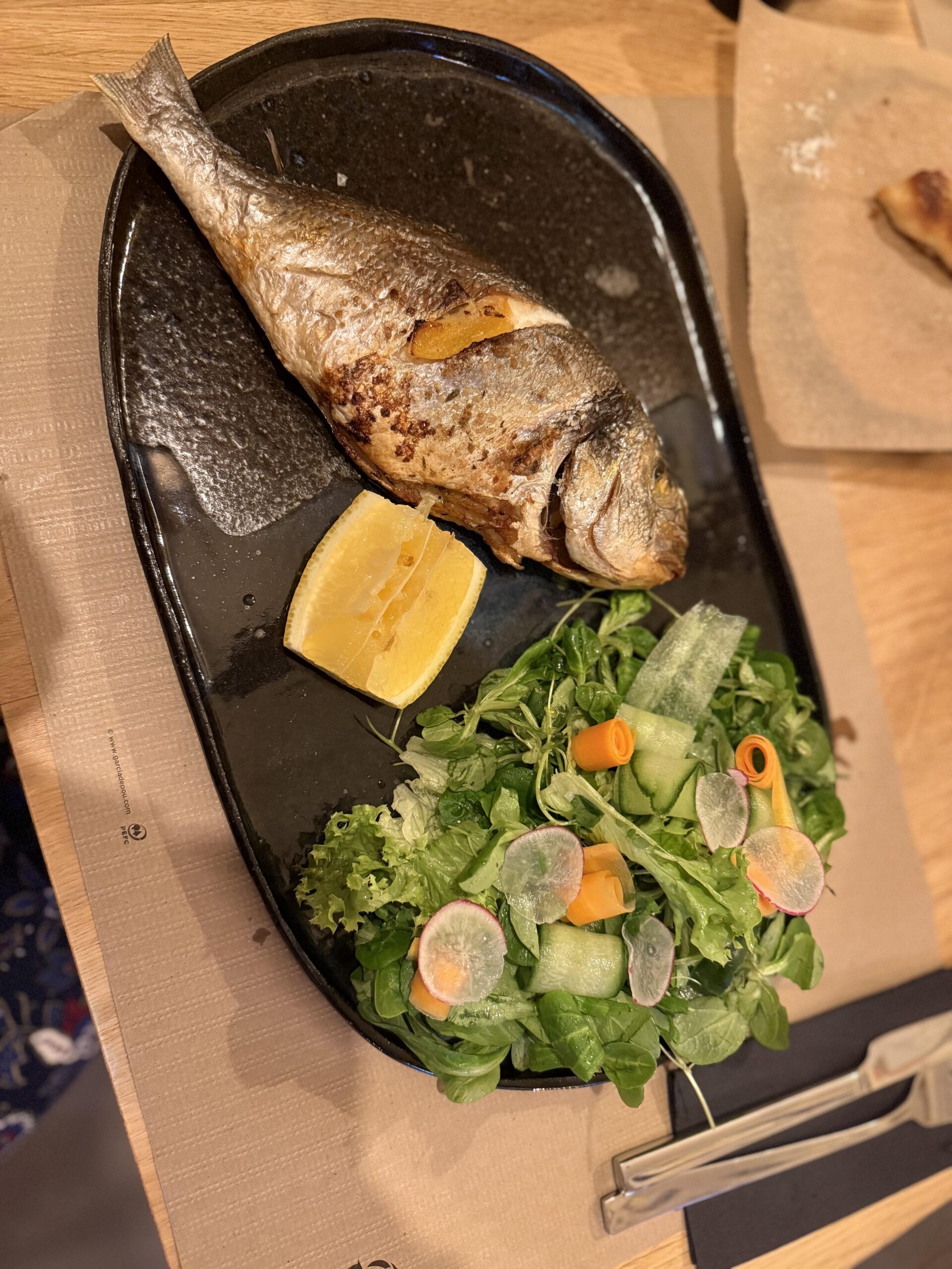 Pieczona dorada podana z cytryną i świeżą sałatką z warzywami na czarnym półmisku.
