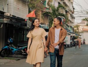 Para (kobieta i mężczyzna) trzyma sie za ręce idąc po ulicy. Oboje mają smart casual strój w stonowanych kolorach.